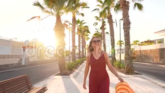 日落时分戴着一顶黄色大帽子的幸福女人沿着棕榈巷散步慢动作视频的预览图