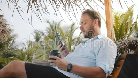 成功的快乐商人喜欢在夏日海滩的躺椅上喝酒思考使用智能手机移动办公应用程序视频的预览图
