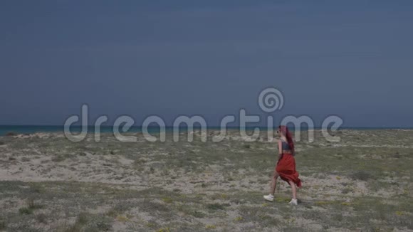 慢动作红发年轻女子穿着红色裙子在蓝天的映衬下沿着海岸走向大海视频的预览图