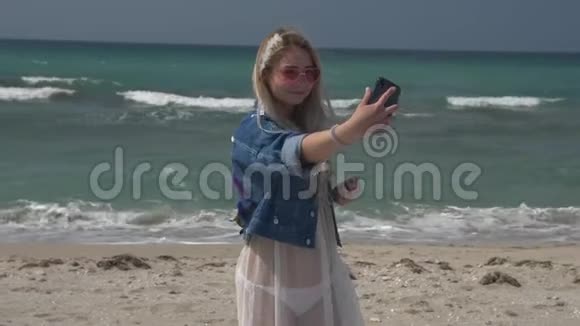 一位身穿半透明白色连衣裙和牛仔夹克的年轻金发女子在电话中以大海为背景拍摄了一张照片视频的预览图