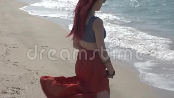 慢动作红头发的年轻女子穿着红色裙子站在海滩上看着海浪视频的预览图