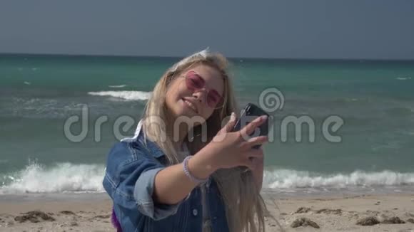 年轻的金发女人穿着一件牛仔夹克在大海的背景下通过视频通话在智能手机上播放她的头发视频的预览图