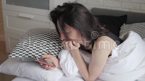 一个幸福的女人躺在她可爱的卧室里躺在白色的床上在智能手机上发短信技术互联网视频的预览图
