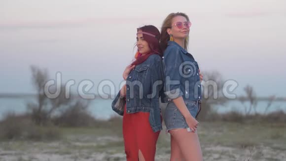 两个穿着牛仔夹克的年轻女子背靠背站在夕阳的柔和光线中头发在夕阳中飞舞视频的预览图