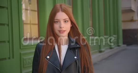 穿着皮夹克走着漂亮的生姜女孩停在镜头前在绿色的房子背景上微笑着走进它视频的预览图