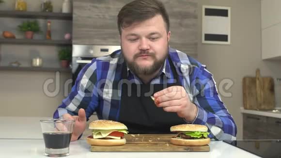 肥胖的白种人坐在厨房里吃快餐汉堡和芝士汉堡喝含有有害苏打水的柠檬水视频的预览图