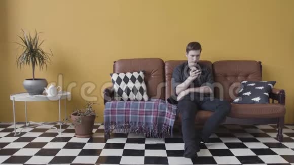 年轻人在黑白地板的房间里坐在一张皮沙发上用手机发送信息视频的预览图