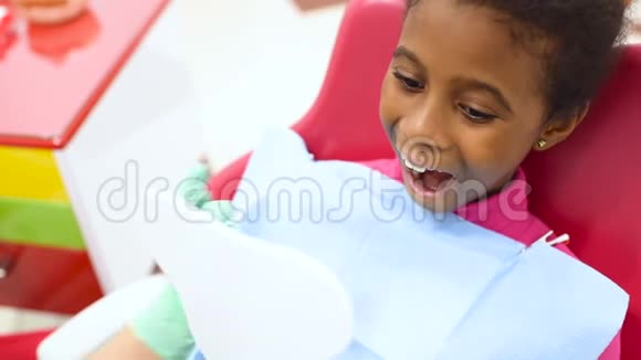 儿童牙医在一张红色和黄色的牙科椅上检查一个可爱的非裔美国女孩的牙齿视频的预览图