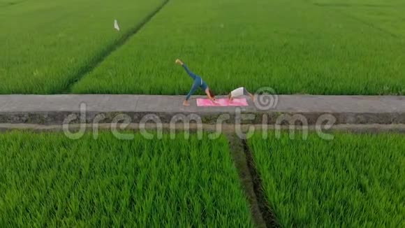 一位年轻女子瑜伽教练在日落时在一个美丽的大稻田上教小男孩的空中拍摄旅费视频的预览图