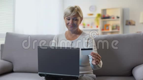 成熟的金发女性在笔记本电脑沙发上输入信用卡号码付款视频的预览图