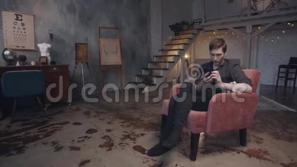 迷人的年轻人坐在舒适的红色扶手椅上智能手机在美丽的黑暗工作室装饰视频的预览图