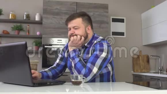 一个留着胡子的男人坐在厨房里手里拿着一台笔记本电脑喝着早间咖啡慢说视频的预览图
