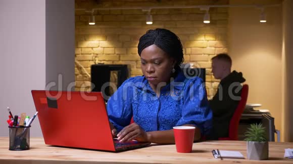 忙碌的非裔美国女商人用笔记本电脑在办公室室内喝咖啡的特写镜头视频的预览图