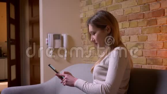在室内沙发上坐着的电话网络上一张年轻迷人的白种人黑发女性浏览网络的特写照片视频的预览图
