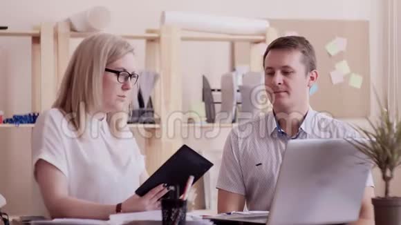 一个女孩和一个白人男子一起在办公室工作一台笔记本电脑和一台平板电脑讨论和记录工作视频的预览图