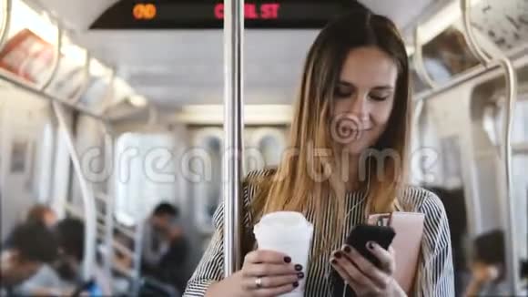 快乐轻松美丽的千禧女孩站在地铁列车上用电子商务程序微笑着看智能手机视频的预览图
