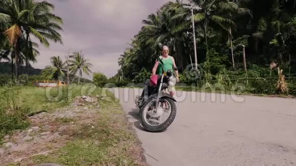 老人坐在摩托车上启动引擎在热带棕榈树景观上骑摩托车的长者视频的预览图