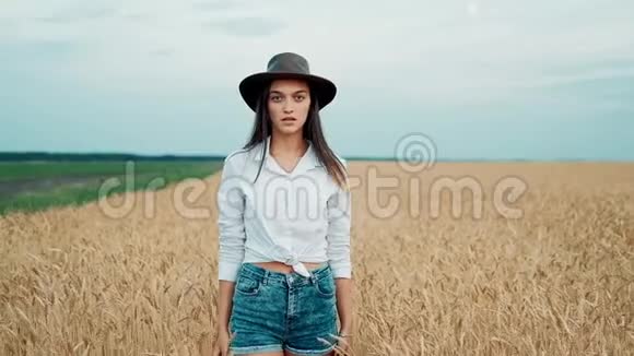 穿着短裤和帽子的年轻女孩走在麦田里在相机里微笑触摸谷物太阳耀斑照耀着视频的预览图