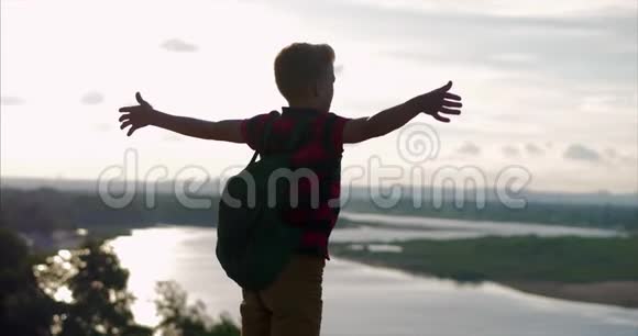 日落时分一个穿着红色衬衫的孩子背上背着一个背包伸手到一座高山上的山顶看着云层视频的预览图