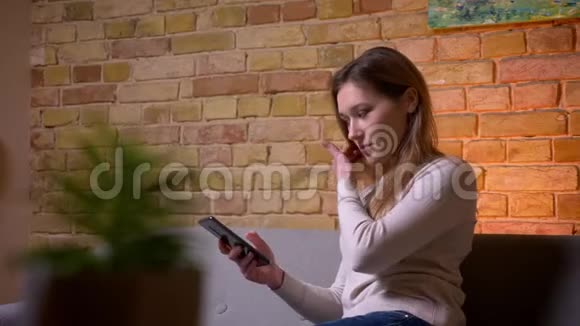 一张年轻迷人的白种人黑发女人的特写照片她在电话里坐在沙发上坐在室内视频的预览图