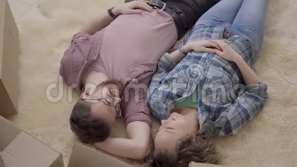 一对年轻夫妇躺在地板上有许多盒子靠近男人和女人说话躺在蓬松的地毯上家庭搬迁视频的预览图