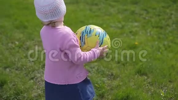 一个两岁的小女孩戴着针织帽手里拿着一个足球球沿着绿草地跑一个慢悠悠的视频的预览图