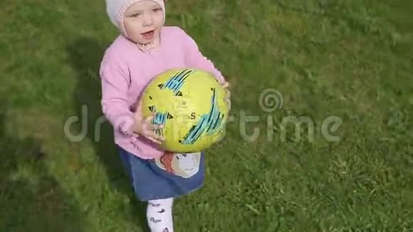 一个两岁的小女孩戴着针织帽手里拿着一个足球球沿着绿草地跑一个慢悠悠的视频的预览图
