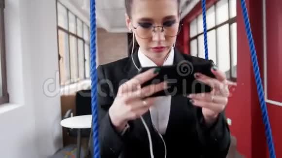 商业概念一个戴眼镜的年轻女子坐在办公室的秋千上听着音乐看着视频的预览图