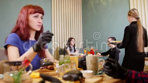 两个戴着黑色手套的人拿着黑色汉堡吃了它看到他们的朋友在另一张桌子旁挥手视频的预览图