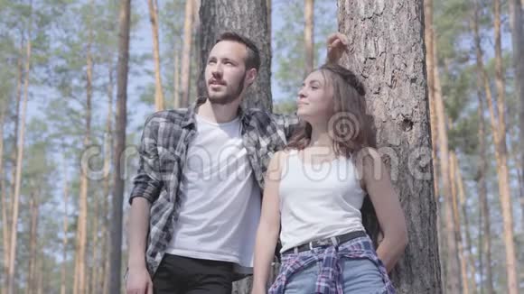 英俊的年轻人和漂亮的女人站在松林里看着远处的肖像露营的概念休闲视频的预览图