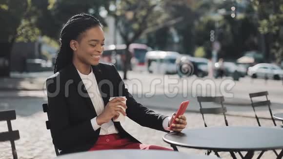 时尚的非洲商务女性使用智能手机喝咖啡坐在舒适的咖啡馆的夏季露台上商务小姐视频的预览图