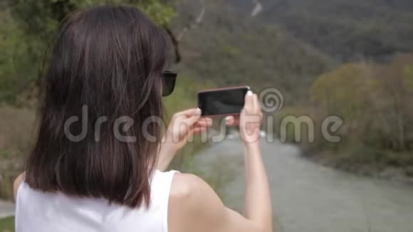 年轻女子在智能手机摄像头上拍摄山河景观照片通过网络社交媒体分享视频的预览图