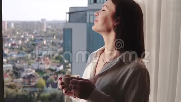 漂亮的深褐色头发的女人享受着她那芳香的咖啡望着窗外从高楼大厦的黄昏小屋视频的预览图