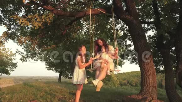 妈妈在树下的秋千上摇着女儿在阳光下玩耍妈妈和宝宝坐在秋千上视频的预览图