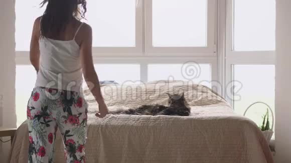 年轻漂亮的女人和她可爱的缅因州浣熊猫躺在床上放松用手机听音乐视频的预览图