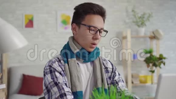 一个穿着衬衫喉咙痛的亚洲年轻人坐在笔记本电脑前的肖像画视频的预览图