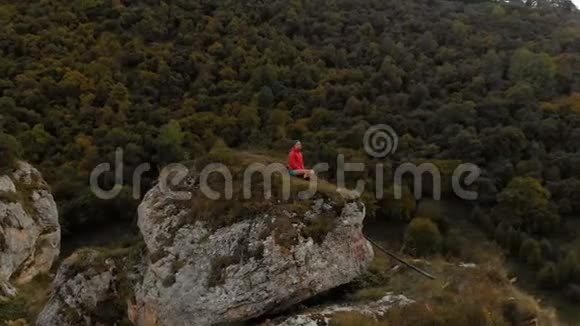 一个年轻人坐在山上岩石上的莲花位置上沉思空中观景四处飞行视频的预览图