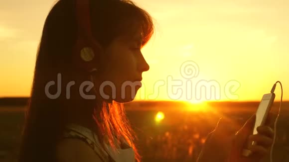 女孩旅行者在智能手机上听音乐在美丽的春天日落的光线下带耳机的年轻女孩视频的预览图