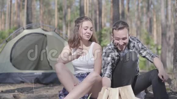 那个年轻人在森林里生火而漂亮的年轻女人坐在附近可爱的情侣在户外休息视频的预览图