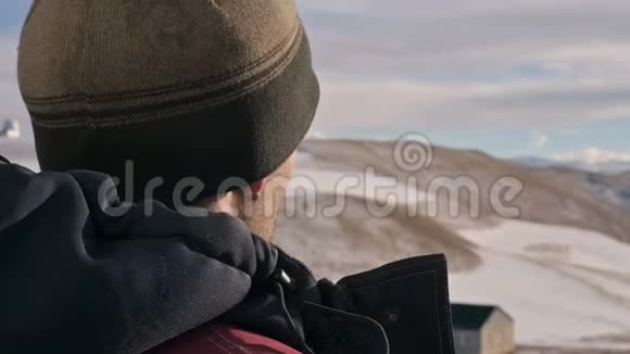 一个穿着温暖夹克和帽子的白人男子在冬天在山上近距离观看群山视频的预览图