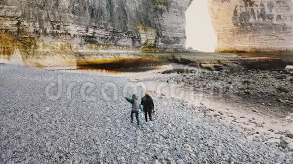 无人机照相机跟随着快乐的游客男人和女人沿着美丽的大卵石海滩走到诺曼底雄伟的岩石视频的预览图