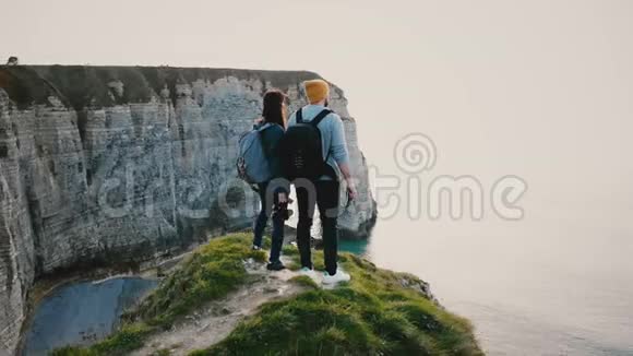 无人机在年轻的快乐旅游夫妇身边飞行背包在诺曼底海岸悬崖上观看史诗般的日落海视频的预览图