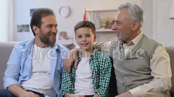 快乐的爸爸和爷爷拥抱着男孩坐在家里的沙发上看着相机视频的预览图