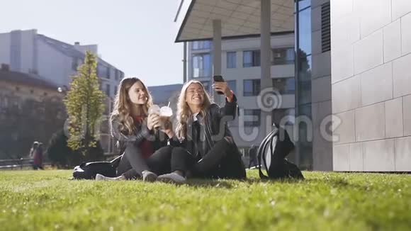 年轻的白种人妇女一边喝着新鲜果汁一边坐在草地上一边用手机自拍照片视频的预览图