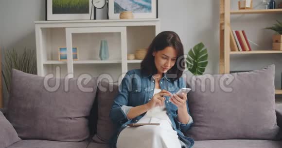 漂亮的女孩在笔记本上写字看着坐在家里的智能手机屏幕视频的预览图