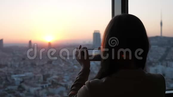 女人在她的现代摩天大楼公寓的智能手机上拍摄日落时的全景城市景色视频的预览图