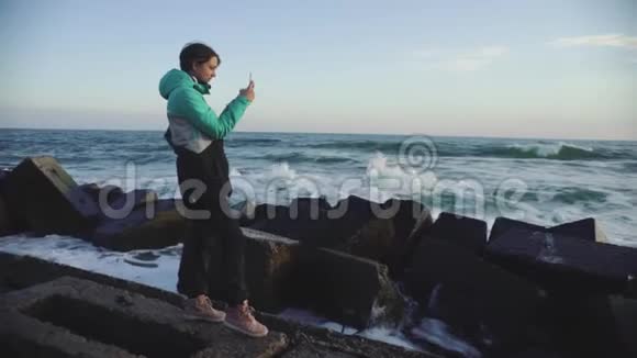迷人的女人穿着温暖的夹克在海边日落时用智能手机拍照慢动作视频的预览图