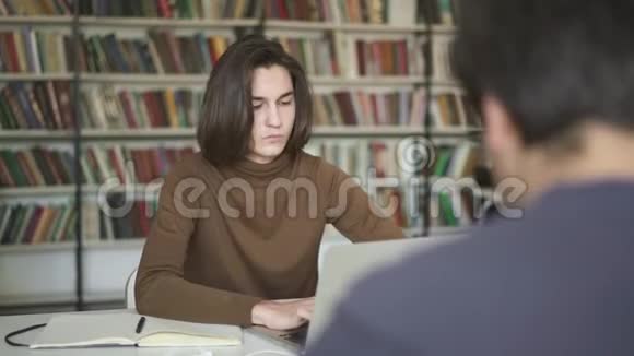学生男子坐在图书馆里用笔记本电脑打字的中间镜头视频的预览图