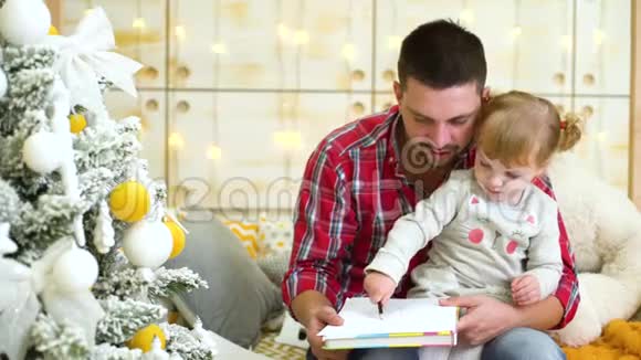 坐在父亲膝上用铅笔在圣诞树旁画画的小女孩视频的预览图