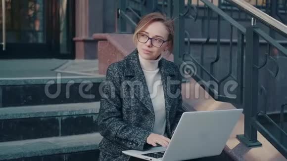 戴眼镜的年轻女人在户外使用笔记本电脑穿外套的女孩坐在台阶上视频的预览图